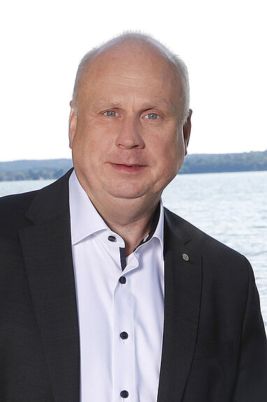 Porträt von Manfred von Dahlen vor Wasser mit Gebäuden im Hintergrund.