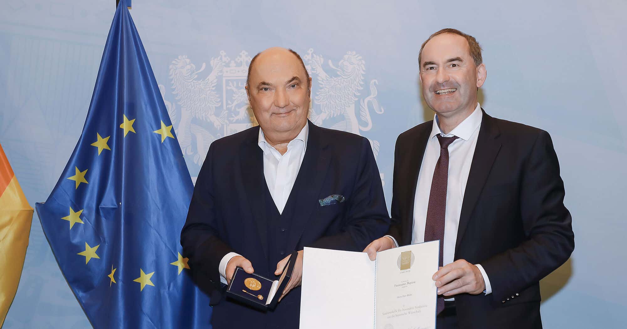Zwei Männer bei der Verleihung der Staatsmedaille, mit Deutschland- und EU-Flaggen im Hintergrund.