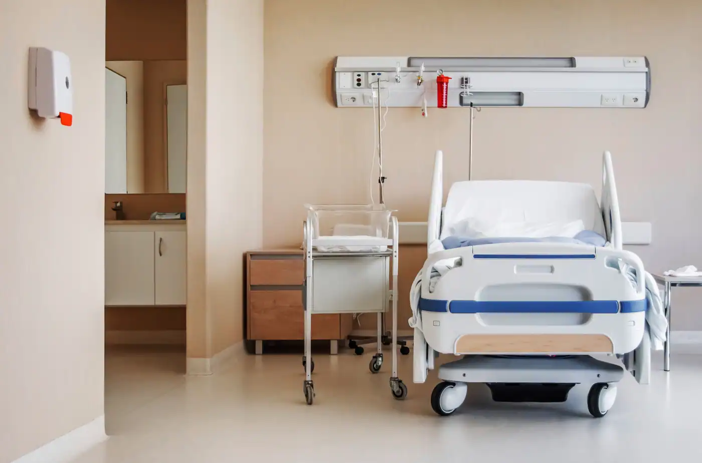 Modernes Krankenhausbett im Patientenzimmer mit Pflegewagen daneben.