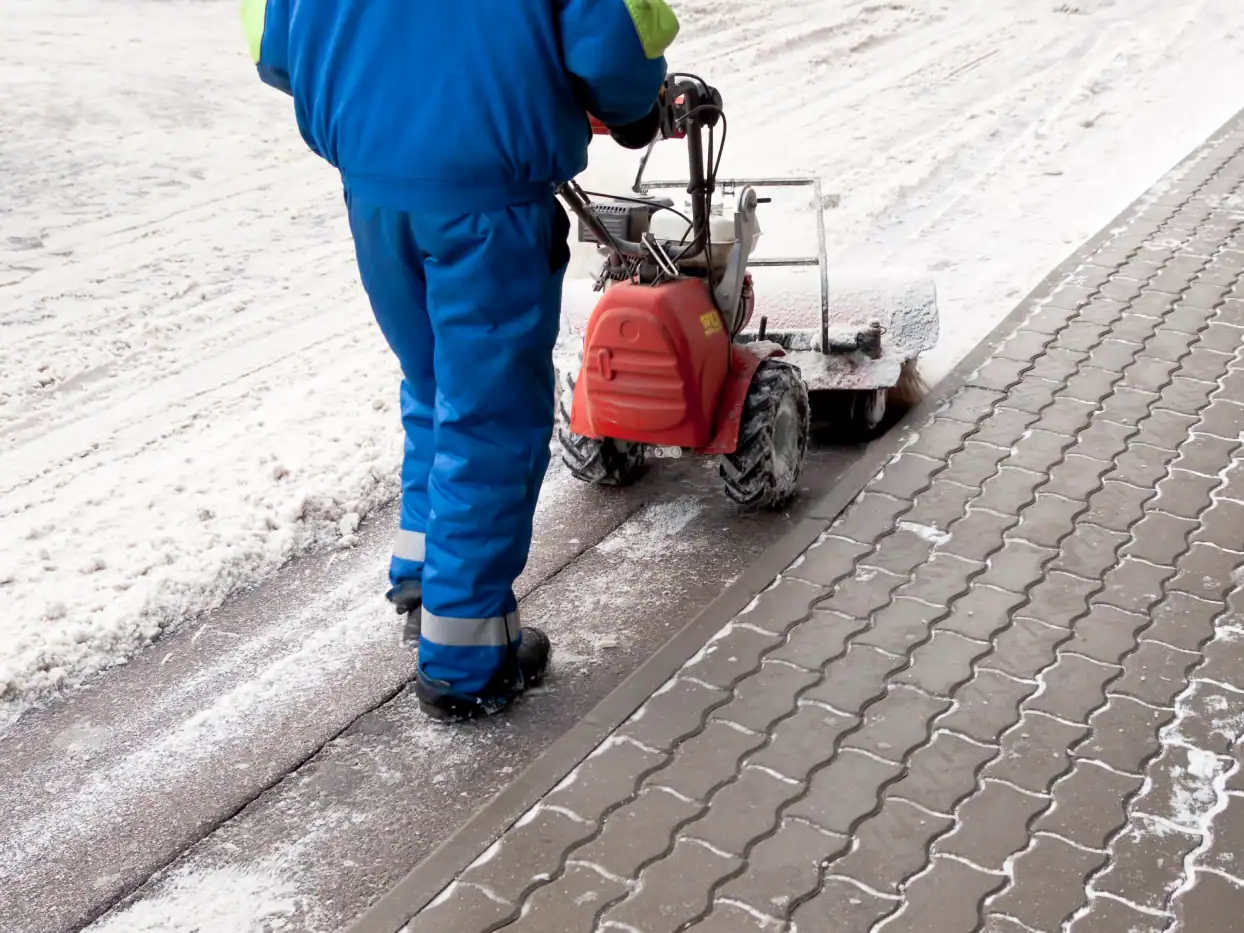 Reinigungskraft entfernt Schnee und Eis vom Gehweg mit Schneeschleuder.