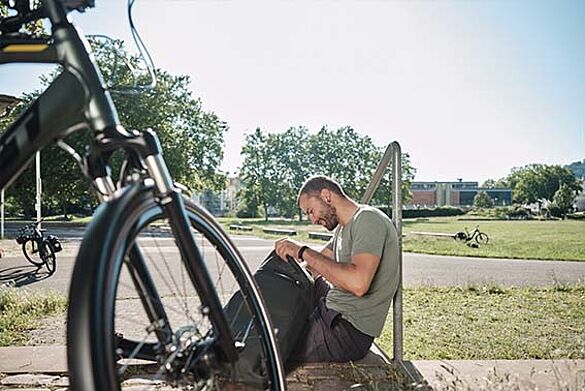 Mann mit Rucksack sitzt im Park, Fahrrad im Vordergrund.